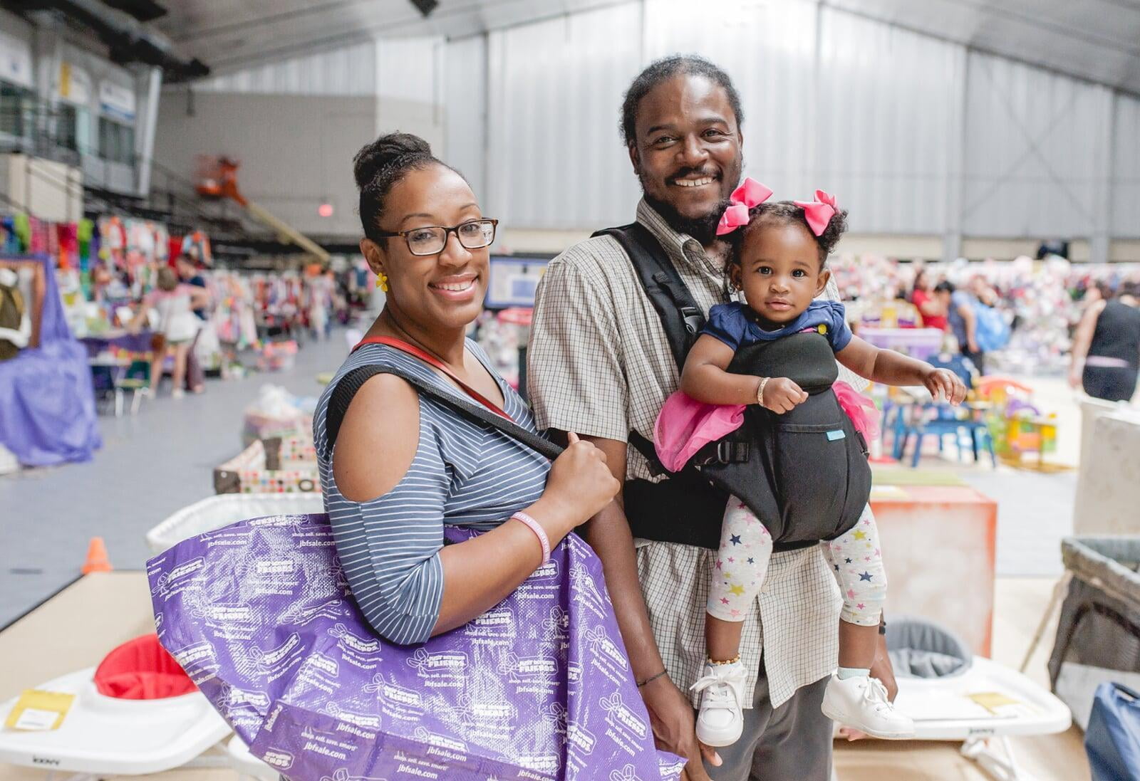 A mom with a large JBF shopping bag on her shoulder stands beside her husband who wears their toddler at a JBF sale.