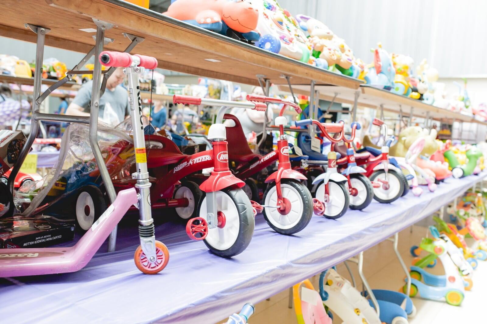 Tricycles, scooters and toys lined up on a table waiting for shoppers to attend the sale.